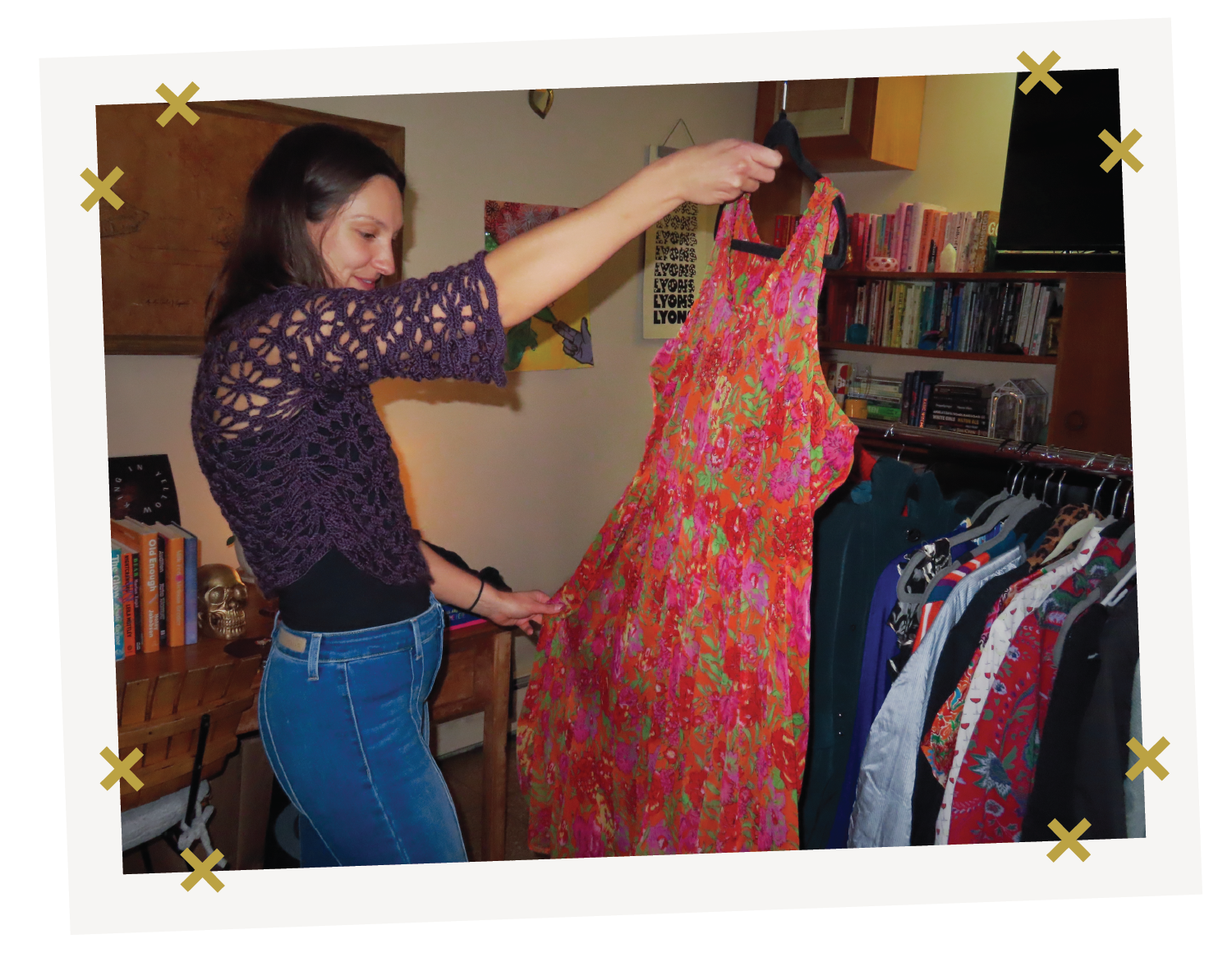 A woman holds up a bright red and pink floral dress at a clothing swap. A rack of secondhand clothes stands behind her.